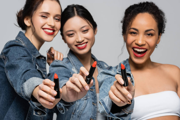 Lápiz labial rojo en manos de alegres mujeres interracial sobre fondo borroso aislado sobre gris  - Foto, imagen