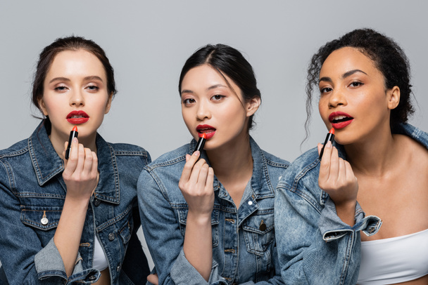 Pretty multicultural women applying red lipsticks and looking at camera isolated on grey  - Foto, imagen