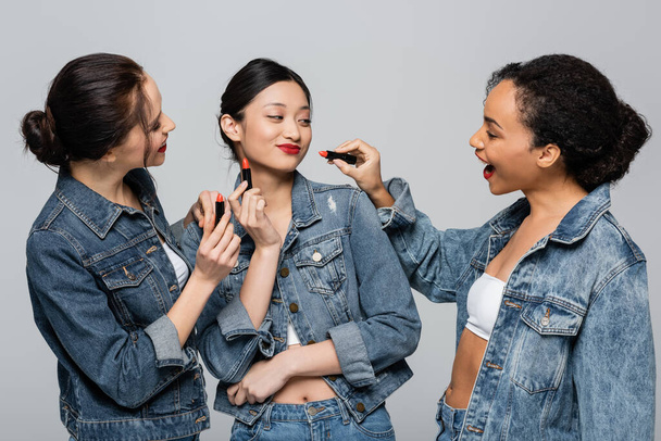 Smiling multiethnic women in denim jackets holding red lipsticks near asian friend isolated on grey  - Foto, immagini