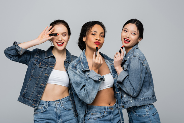 Young interracial women in denim jackets holding red lipsticks isolated on grey  - Photo, Image