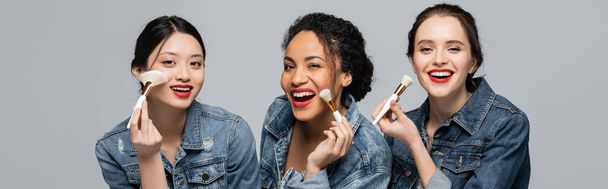 Pretty multiethnic women with red lips holding cosmetic brushes and looking at camera isolated on grey, banner  - Photo, Image