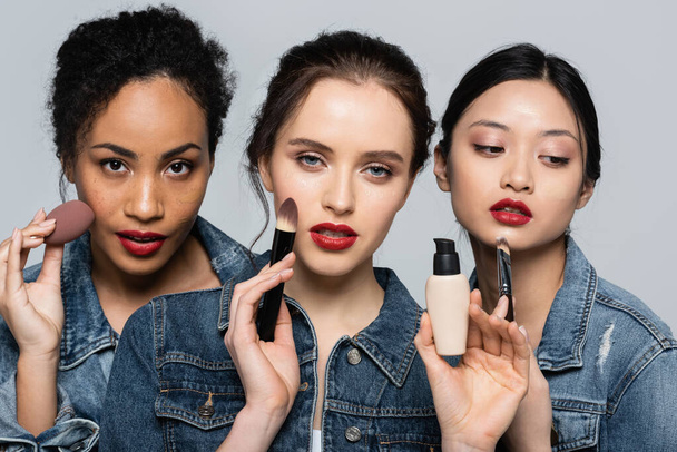 Interracial women with red lips holding cosmetic brushes and face foundation isolated on grey  - Photo, Image