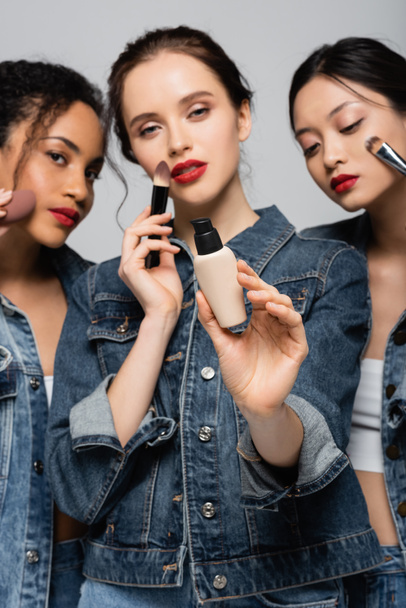 Face foundation in hand of blurred woman near interracial friends with cosmetic brushes isolated on grey  - Фото, изображение