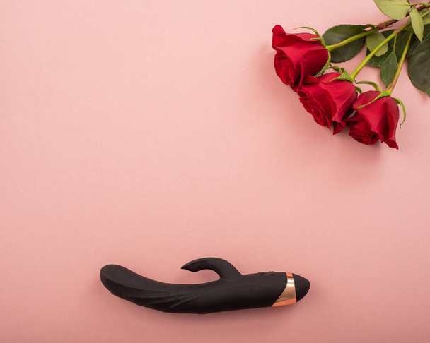 Top view of a black dildo and a bouquet of scarlet roses on a pink background. Copy space - Φωτογραφία, εικόνα