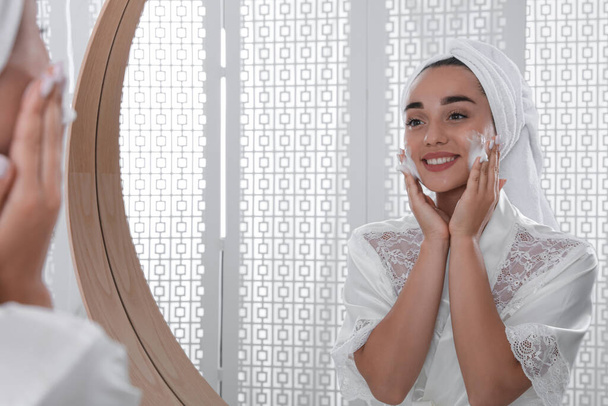 Beautiful young woman applying cleansing foam onto face near mirror in bathroom. Skin care cosmetic - Foto, afbeelding