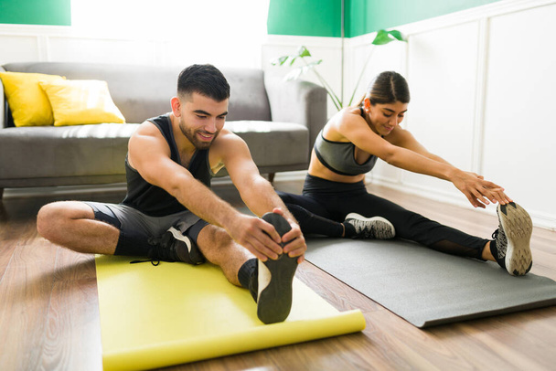 Handsome latin man and happy young woman stretching the legs on an exercise mat in the living room - Фото, изображение