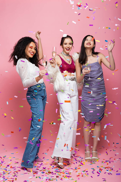 Full length of smiling multiethnic friends with champagne standing under confetti on pink background - Foto, immagini