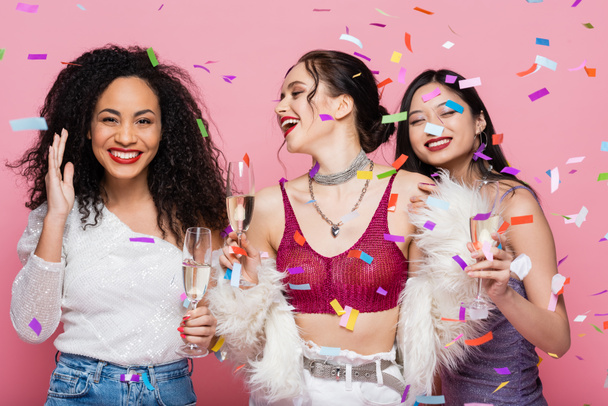 Trendy multiethnic women with champagne standing under confetti on pink background - Foto, Imagem