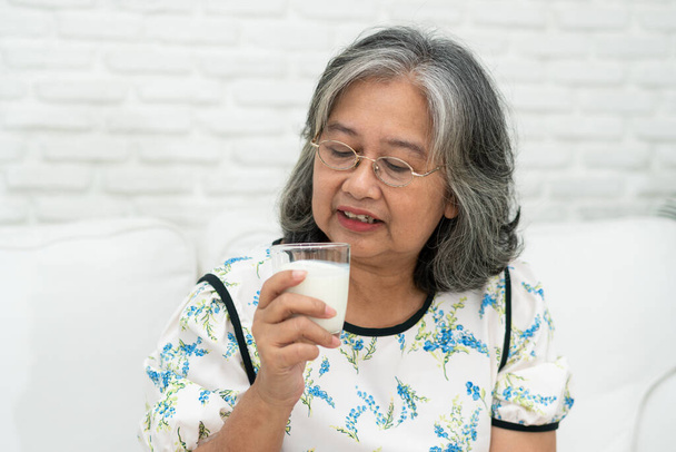 Asiatische Seniorin hält Glasmilch in der Hand, während sie es sich auf einem Sofa im Wohnzimmer für Wellness im Ruhestand gemütlich macht. Ältere Frau trinkt ein Glas Milch, um ihr Wohlbefinden aufrechtzuerhalten. Konzept von Wellness und Gesundheit. - Foto, Bild