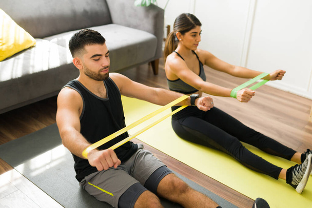 Подходящая пара в спортивной одежде, сидящая на ковриках для упражнений и держа банды сопротивления во время тренировки рук  - Фото, изображение