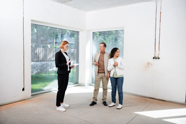 redhead realtor in glasses holding digital tablet near interracial couple inside of new house - Photo, Image