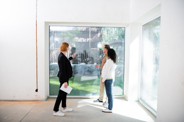 redhead realtor in glasses holding digital tablet while pointing at panoramic window near interracial couple inside of new house - Fotoğraf, Görsel