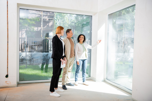 redhead realtor in glasses holding digital tablet near african american woman pointing with hand at panoramic window inside of new house - Valokuva, kuva