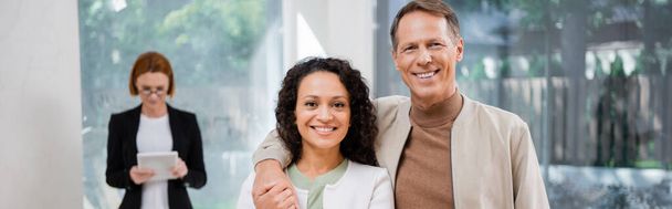 feliz pareja interracial sonriendo cerca pelirroja agente inmobiliario en gafas sosteniendo tableta digital sobre fondo borroso, bandera - Foto, Imagen
