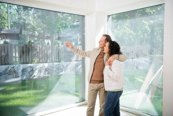 wesoły mężczyzna wskazując ręką na panoramiczne okna przytulając żonę w nowym domu - Zdjęcie, obraz