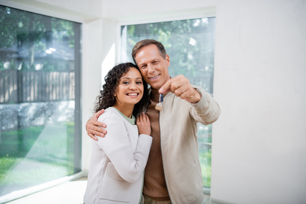 cheerful man holding blurred key and hugging african american wife in new house - Photo, Image