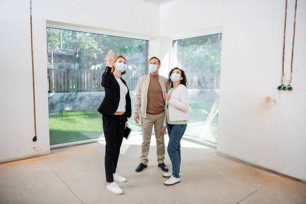 異人種間のカップルの近くに立っている間に新しい家で手で指差す医療マスクの赤毛の不動産業者  - 写真・画像