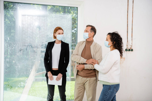 redhead realtor in medical mask holding digital tablet and looking at interracial clients  - Fotografie, Obrázek