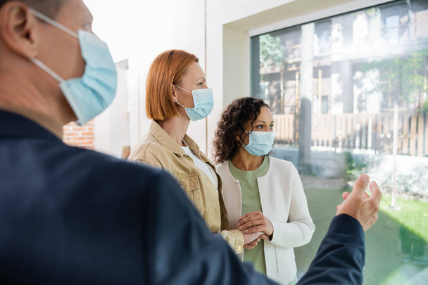 blurred realtor in medical mask pointing at panoramic window near interracial lesbian couple inside of new house - Fotoğraf, Görsel