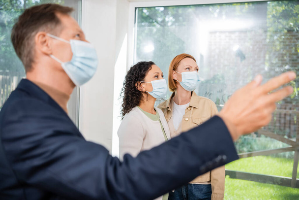 blurred realtor in medical mask pointing with hand near interracial lesbian couple inside of new house - Fotografie, Obrázek