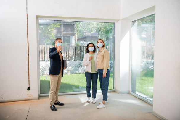 agent immobilier d'âge moyen dans le masque médical pointant avec la main près de couple lesbien interracial à l'intérieur de la nouvelle maison - Photo, image