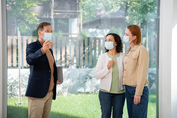middle aged realtor pointing with hand near interracial lesbian couple in medical masks inside of new house - Фото, изображение