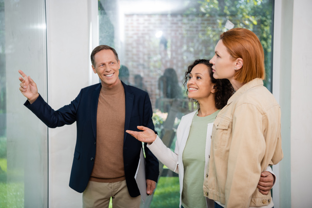 happy realtor holding folder and pointing at panoramic window near interracial lesbian couple inside of new house - Photo, Image