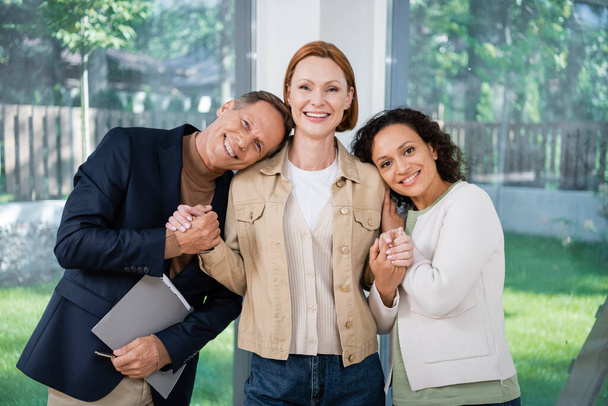 happy realtor holding hands with cheerful and interracial lesbian couple  - Photo, Image