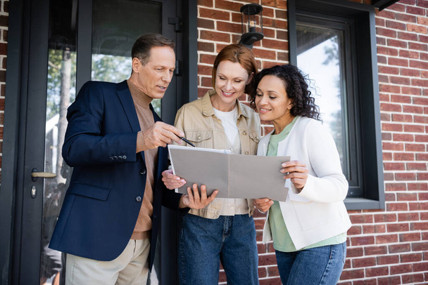middle aged realtor holding folder near happy and interracial lesbian couple  - Foto, Imagen