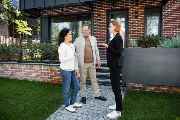 realtor in glasses gesturing while talking with african american woman near husband and modern house - Photo, Image