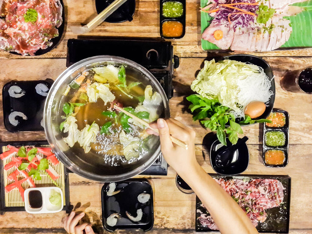mão segurando pauzinhos comendo.Sukiyaki e sopa de shabu com Dumplings, repolho branco, caranguejo, vermicelli, camarão, e em uma xícara branca em madeira - Foto, Imagem