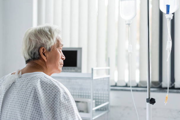Visão lateral de idosos asiático paciente olhando para terapia intravenosa no hospital  - Foto, Imagem