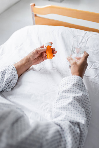 Oříznutý pohled na staršího pacienta držícího vodu a pilulky na lůžku na klinice  - Fotografie, Obrázek