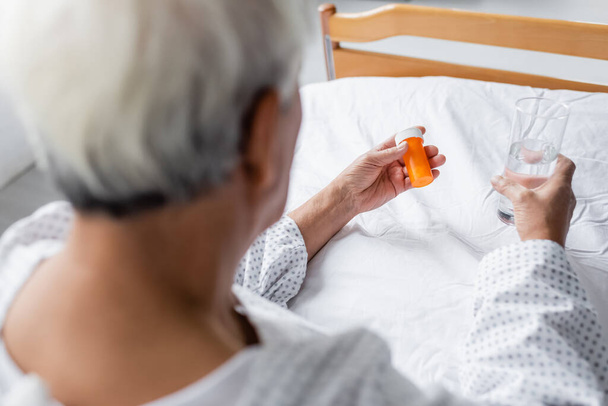 Blurred elderly patient holding pills and glass of water on bed in hospital  - Zdjęcie, obraz