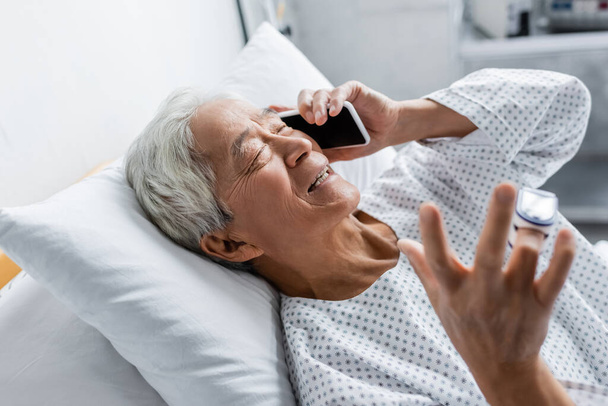 Uśmiechnięty azjatycki pacjent z pulsoksymetrem rozmawiający na smartfonie w klinice  - Zdjęcie, obraz