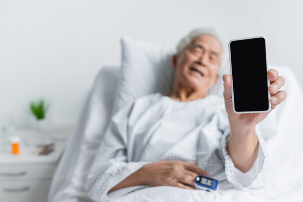 Smartphone στο χέρι του θολό ασιατικό ασθενή με παλμικό οξύμετρο στο θάλαμο του νοσοκομείου  - Φωτογραφία, εικόνα
