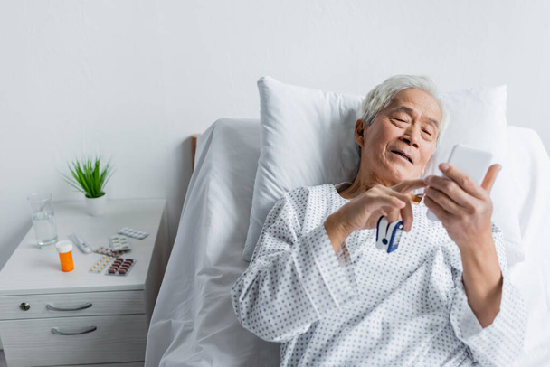 Ηλικιωμένος Ασιάτης ασθενής με παλμικό οξύμετρο χρησιμοποιώντας smartphone στο κρεβάτι κοντά σε χάπια και νερό στην κλινική  - Φωτογραφία, εικόνα