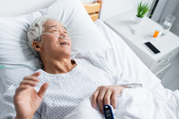 Vista ad alto angolo di sorridente paziente asiatico con pulsossimetro e cannula nasale sdraiato sul letto in clinica  - Foto, immagini