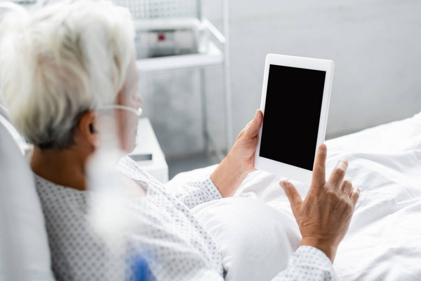 Θολή ασιατική ασθενή με ρινική κάνουλα κρατώντας ψηφιακό δισκίο με κενή οθόνη στο κρεβάτι στην κλινική  - Φωτογραφία, εικόνα