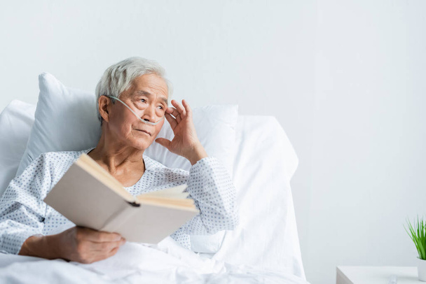 Asiática paciente en vestido y cánula nasal celebración libro en la cama en la clínica  - Foto, Imagen
