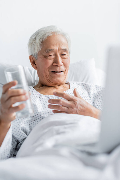 Usmívající se asijský pacient drží sklenici vody v blízkosti rozmazané notebook na nemocniční posteli  - Fotografie, Obrázek