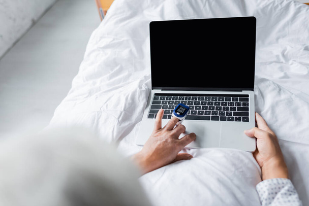 Nabız oksimetresi olan hastanın klinikte yatağında boş ekranla dizüstü bilgisayarı kullanmasının yüksek açılı görüntüsü.  - Fotoğraf, Görsel