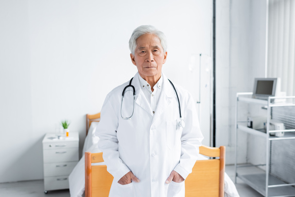 Ázsiai orvos fehér köpenyben nézi a kamerát a kórházi kórteremben  - Fotó, kép