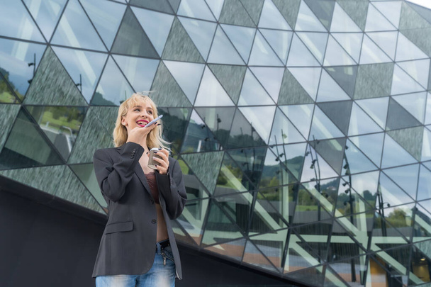 Entrepreneurial girl business park in a modern building with colored glass, young blonde woman in suit sending a voice note - Zdjęcie, obraz