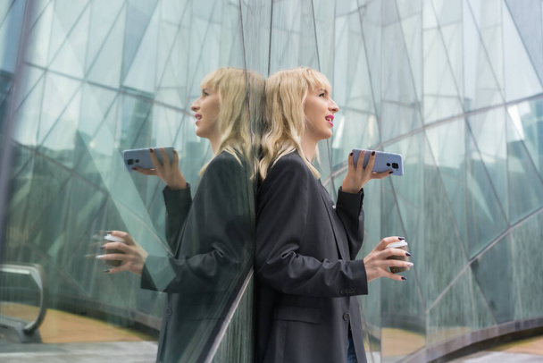 Entrepreneurial girl business park in a modern building reflected in the windows, young blonde in a suit sending a voice message with a take away coffee - Φωτογραφία, εικόνα