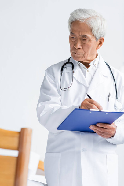 Elderly asian doctor in white coat writing on clipboard in hospital ward  - Φωτογραφία, εικόνα