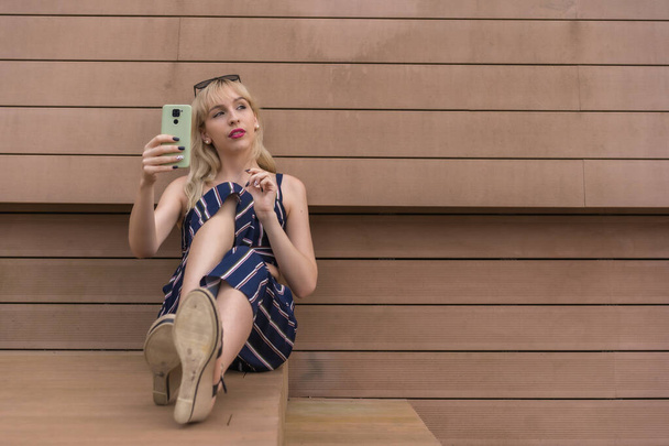 Entrepreneur girl sitting in a business park on a wooden wall, young blonde taking a selfie with the phone, copy space and paste - Photo, Image