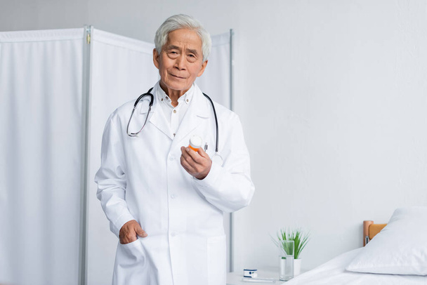 Senior asian doctor holding pills and looking at camera in clinic  - Φωτογραφία, εικόνα