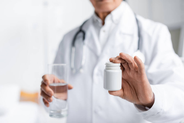 Cropped view of blurred doctor holding jar with pills and glass of water in clinic  - Фото, изображение