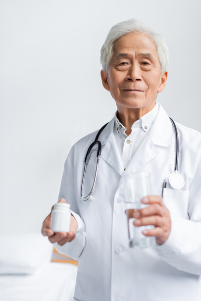 Senior asian doctor holding glass of water and jar with pills in hospital ward  - 写真・画像